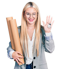 Wall Mural - Beautiful blonde woman holding paper blueprints and house keys winking looking at the camera with sexy expression, cheerful and happy face.