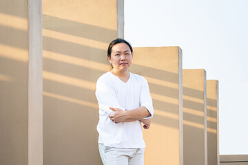 Asian long hair, Man in white long arm t-shirt is standing at the rooftop outdoor field with light brown wall bacgkround.