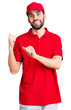 © Krakenimages.com - Young handsome man with beard wearing delivery uniform pointing to the back behind with hand and thumbs up, smiling confident