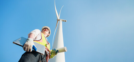Poster - Low angle view of Architectural engineer working with his laptop and blueprints in hand while supervising construction site in Wind turbine farm power generator in beautiful nature landscape.