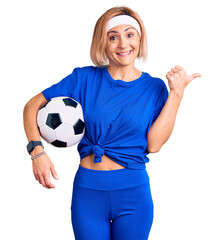 Sticker - Young blonde woman holding soccer ball pointing thumb up to the side smiling happy with open mouth