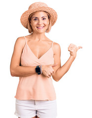 Canvas Print - Young blonde woman wearing summer hat pointing to the back behind with hand and thumbs up, smiling confident