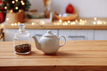 A Ceramic White Teapot And A Transparent Glass Jar With Tea Leaves Stand On A Wooden Table In The Background Of The Kitchen, Glowing Garlands And A Christmas Tree. Family Breakfast At Home.