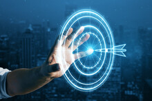 Target Concept With A Glowing Hologram Of Bullseye And Arrow Hitting The Center. Goals Achievement, Success. Close Up Of Businessman Hand Using Hud Screen On Blurry Night City Background.