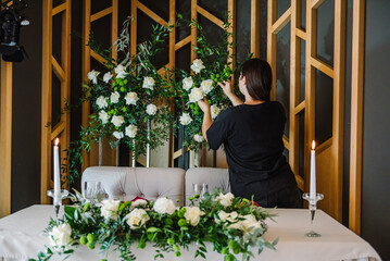 Organizer women arranges and decoration wedding. Setup. Table newlyweds in banquet hall on party. Festive table with arch, decorated with composition of flowers and greenery, candles. Back view.