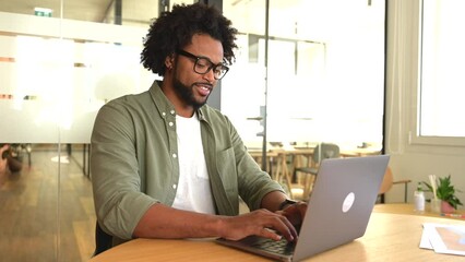 Focused curly handsome man using laptop computer in contemporary coworking space, freelance programmer in glasses coding sitting at the office desk, creativity designer or architect typing