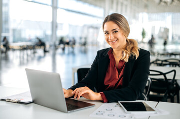 Photo of a confident lovely elegantly dressed positive caucasian business lady, executive, recruitment, product manager, sits in a business center, working in a laptop, looks at camera, smile friendly