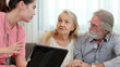 © Benixs - Caucasian young adult nurse is visiting elderly couple and holding a tablet to show medical records. Female caregiver is explaining result of medical examination and treatment to senior grandparents. 