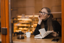 Woman Wear Black Sweater And Eye Glasses, Reading A Paper Book With A Cup Of Tea In Outdoor Cafe, Hobby Or Leisure. Happy Smiling Woman Read And Look Dreaming And Thoughtful. Girl Flip Pages.