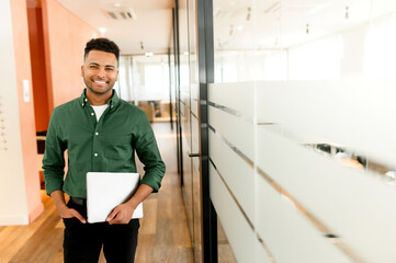 Inspired young indian businessman looks at camera and smiles cheerfully, standing in modern office hallway and holding laptop. Male office employee, freelancer, designer full of new ideas and creative