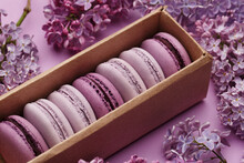 Set Of Violet And Lilac Macaroons In Cardboard Gift Box With Lilac Flowers On Violet Background. Close Up View, Selective Focus. Traditional French Dessert