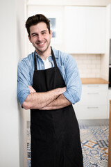 handsome young man smiling in the kitchen wearing an apron - Portrait of a cheerful confident caucasian young man looking at camera with arms crossed 