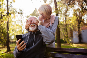 Wall Mural - Active modern cheerful senior couple meet in the park. A older mar is sitting on a park bench using smartphone, and an elderly woman surprises him from the back.