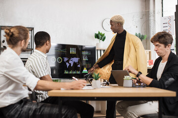 Wall Mural - Group of four multi ethnic partners sitting at desk and listening speech of african american man near huge monitor with various graphs and charts. Business meeting at office.