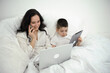 © Oleksandra - work at home online mom and son sit in bed under a white blanket in their hands laptop tablet phone lessons online work on the internet woman talking on the phone smiling making order solves business