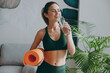 © ViDi Studio - Young strong sporty athletic fitness trainer instructor woman wearing green tracksuit holding in hand yoga mat drink water training do exercises at home gym indoor. Workout sport motivation concept.