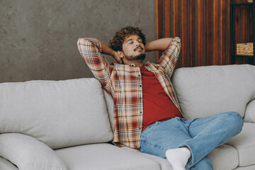 Wall Mural - Young calm Indian man wearing casual clothes hold hands behind neck close eyes sits on grey sofa couch stay at home hotel flat rest relax spend free spare time in living room indoor. Lounge concept.