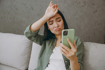 Close up sad young woman of wear casual clothes hold in hand use mobile cell phone put hand head sits on grey sofa couch stay at home hotel flat rest relax spend free spare time in living room indoor