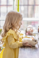 Wall Mural - A little blonde girl in a yellow dress is sitting at a festive Easter table with rabbits.A baby and a rabbit. The concept of Easter.Easter at the table. The Christian tradition.