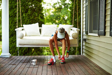 A Female Runner Laces Up Her Running Shoes On Her Porch Swing.