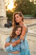 © svetograph - Mother daughter sunset. in blue dresses with flowing long hair against the backdrop of sunset. The woman hugs and presses the girl to her.