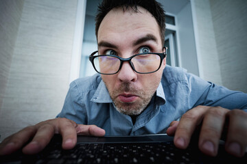 The emotion of surprise on the face. The astonished worker looks at the screen. young handsome man in a blue shirt is working at a computer on a light background. The programmer is focused on work.