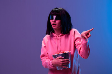 Wall Mural - Excited awesome brunet woman in pink hoodie trendy specular sunglasses with popcorn point finger aside posing isolated in blue violet color light background. Neon party Cinema concept. Copy space