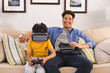 © Wavebreak Media - Portrait of hispanic father sitting by son playing video games through vr headset in living room