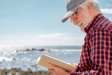 Wall Mural - Portrait of handsome senior bearded man reading a book in front to the sea. Elderly relaxed pensioner with eyeglasses and hat enjoying retirement in outdoor