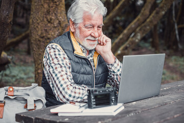 Sticker - Handsome smiling senior man working on laptop while sitting at the wooden table outdoors in the middle of the park. Concept of smart working in nature, business in fresh air