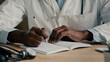 © Yuliia - Unknown male hands african man doctor sit at clinic desk writing medical prescription prescribe hospital referral filling checklist health symptoms write insurance make notes in registration notebook