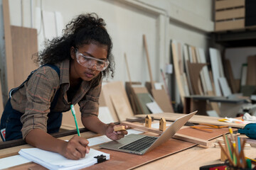 Young female carpenter working in wood workshop. Black female carpenter wearing safety glasses working in workshop at wooden warehouse. SME, Start up and small business concept