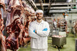 A confident meat factory worker is standing by the big pieces of meat in slaughter house.