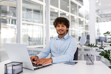 Portrait of young programmer inside office with laptop, indian man smiling and looking at camera, man typing on keyboard, writing code for software, happy with achievement results.