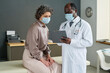 © pressmaster - Experienced African American male general practitioner in lab coat and protective mask explaining prescriptions to female patient