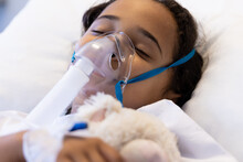 Sick Biracial Girl Sleeping With Oxygen Mask And Teddy Bear On Hospital Bed