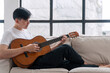 © kaew6566 - Happy asian handsome man playing acoustic guitar sitting on sofa relax beside window at apartment. Young male enjoying play string music with acoustic guitar hobby in living room.Leisure hobby concept