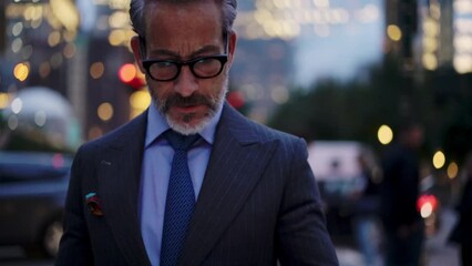 Serious mature successful man in suit and stylish eye glasses typing text message on mobile phone device while walking on city street at evening. Businessman browsing network on cellular application