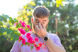 Portrait of handsome young allergic man is suffering from pollen allergy or cold on natural flowers, flowering tree background at spring or sunny summer day, sneezes, blowing his runny nose rubs eyes