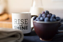 A Mug With Rise And Shine On It Sits Among Breakfast Items Of Blueberries And Milk.