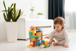 © stenkovlad - Baby girl playing indoors sitting on floor in playroom at home with constructor. Educational game for baby and toddler. Children build toy house. Creative