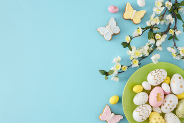 Wall Mural - Easter decoration concept. Top view photo of green plate with colorful easter eggs butterfly shaped gingerbread and cherry blossom branch on isolated pastel blue background with empty space