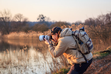 photographer with camera, telephoto lens and backpack taking picture at lake during sunset. hobby an
