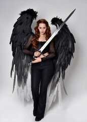 Canvas Print - Full length portrait of beautiful woman with red hair wearing corset, leather pants, large black angel feather wings. Standing walking pose holding sword weapon. Isolated on white studio background.