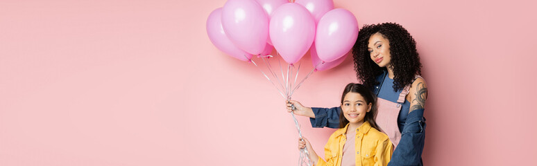 Tattooed woman holding balloons near smiling child on pink background, banner.