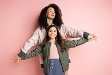 Curly Mother Holding Hands Of Cheerful Daughter On Pink Background.