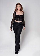 Canvas Print - Full length portrait of beautiful woman with long red hair wearing black corset top and leather pants. Standing pose, walking forwards with gestural hands reaching out. Isolated on white studio backgr