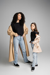 Preteen child in trench coat posing near stylish mom on grey background.