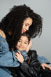 © LIGHTFIELD STUDIOS - Curly mother hugging smiling daughter in leather jacket isolated on grey.
