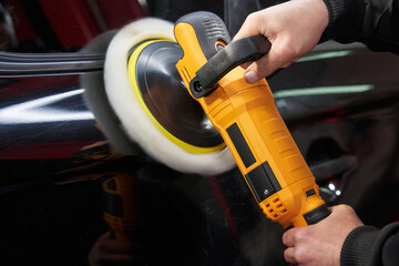 Wall Mural - caucasian worker's hands hold the handles of a yellow angle grinder with a polishing wheel and polish the paintwork of a black car